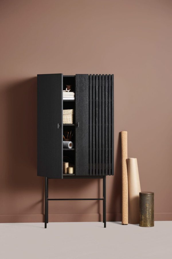 Et moderne, sort træskab med en dør åben, afslørende hylder med pænt arrangerede ting, står på en blød beige baggrund. Ved siden af læner tre cylindriske genstande op ad væggen. Dette stykke indkapsler
