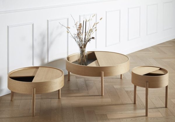 Tre moderne indlejrede sofaborde af lyst træ, med cirkulære plader og tynde ben. Det største bord rummer en vase med tørrede blomster. Disse er sat på et trægulv mod en hvid panelvæg