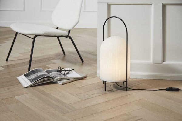 Et moderne værelse med en minimalistisk gulvlampe med et matteret glascover ved siden af et åbent magasin og et par briller på et sildebenstrægulv. En hvid stol med stofsæde