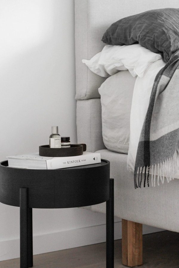 Et monokromt soveværelseshjørne med en WOUD - Arc Sidebord Ø42 med bøger, en flaske og et hyggeligt grå- og hvidstribet tæppe draperet over en let-tonet minimalistisk designermøbel seng.
