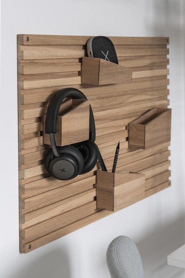 En moderne træorganisator designet af Tomorrowsdesign, monteret på en hvid væg med slidser og en krog, der holder hovedtelefoner, en konvolut og småting som kuglepenne og en notesbog.