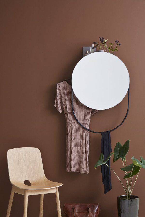Et minimalistisk boligindretningsarrangement med et rundt hvidt spejl på en brun væg, en beige designerstol, en potteplante og hængende beklædningsgenstande. En lille hylde over spejlet viser en plante.