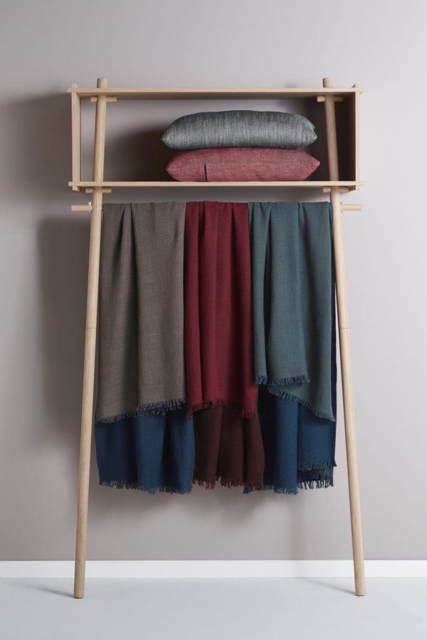 A WOUD - Töjbox Garderobe med en grå pude på toppen, der holder adskillige tørklæder i nuancer af blå, rød og grå, alle draperet pænt over den vandrette stang mod en neutral baggrund. Dette stykke fordobles