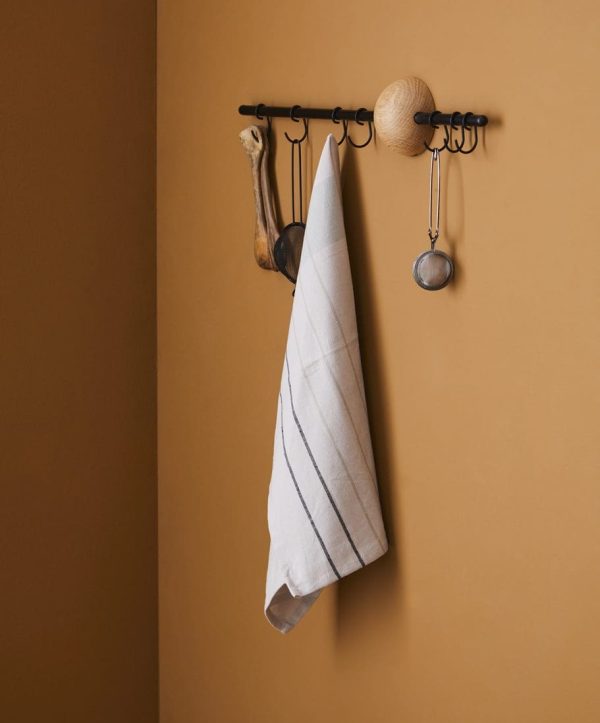 Et pænt foldet køkkenhåndklæde hænger fra en sort metalreol designet af Tomorrowsdesign på en varm beige væg, ledsaget af køkkenredskaber inklusive en øse og en si.