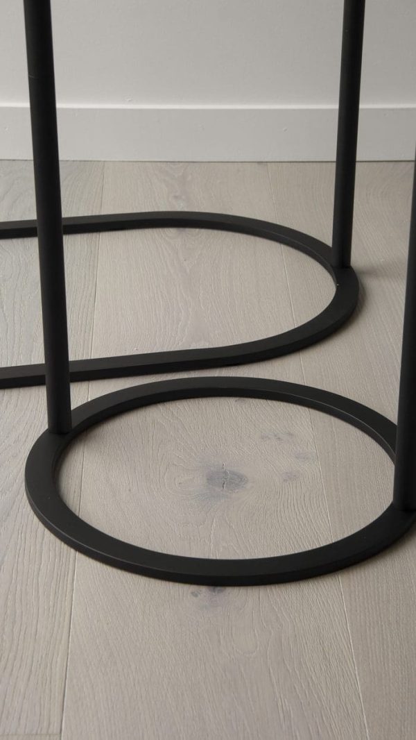 Et nærbillede af et lyst trægulv med de sorte metalben på en skammel, der viser cirkulære fodstøtter, der krydser hinanden geometrisk. Dette designermøbel-element fremhæver ethvert interiør med sit