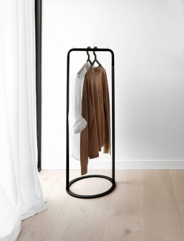 Et minimalistisk tøjstativ med sort ramme på et trægulv, med en brun sweater og en hvid beklædningsgenstand. Stativet, der er placeret nær et hvidt gardin i et rum med stærkt naturligt lys, er en
