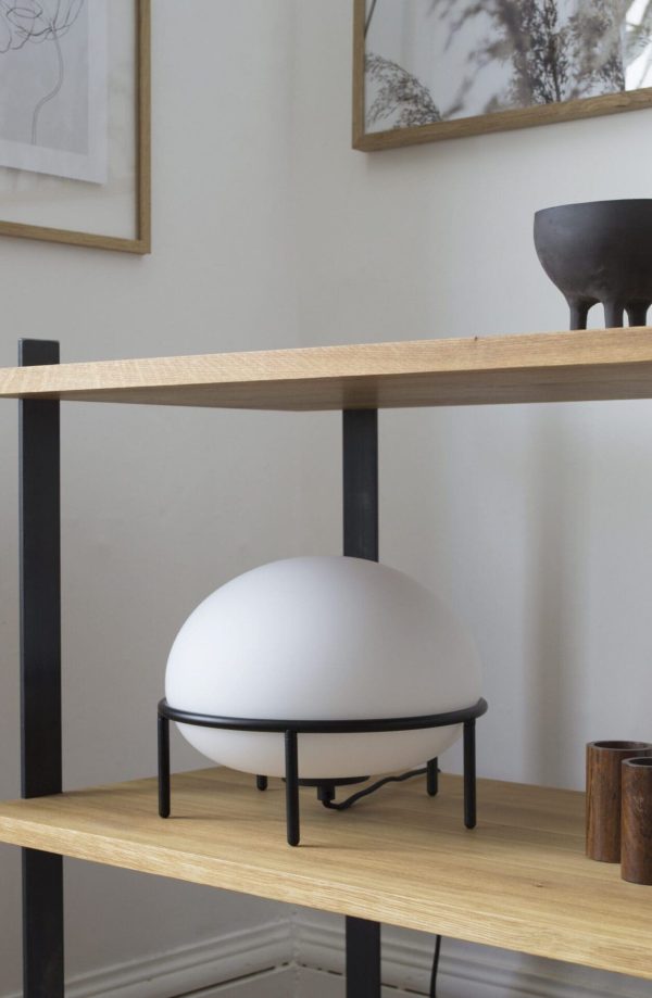 Et minimalistisk værelse med en træhylde med sorte metalrammer, der viser en WOUD - Pump Bordlampe på et lavt stativ, en sort skål og forskellige trægenstande. Der er designerindrammet kunstværk ovenfor.