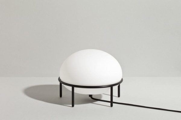 En minimalistisk hvid oval-formet lampe på et slankt sort metalstativ, med et Tomorrows-design-æstetik, med en synlig strømledning, der strækker sig ud fra den, placeret mod en almindelig lysegrå baggrund. WOUD - Pump Bordlampe.