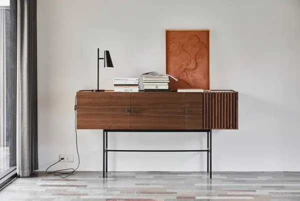 Et minimalistisk værelse med en træcredenza med bøger og en lampe på toppen, placeret mod en lysegrå væg ved siden af et gulv-til-loft-vindue. Et abstrakt rødt kunstværk af Tomorrowsdesign hænger
