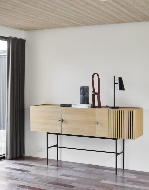 Et moderne minimalistisk værelse med en træskænk på en tynd metalramme, sat mod en almindelig væg ved et vindue med mørke gardiner. Værelset har møbler som et træloft og en sild