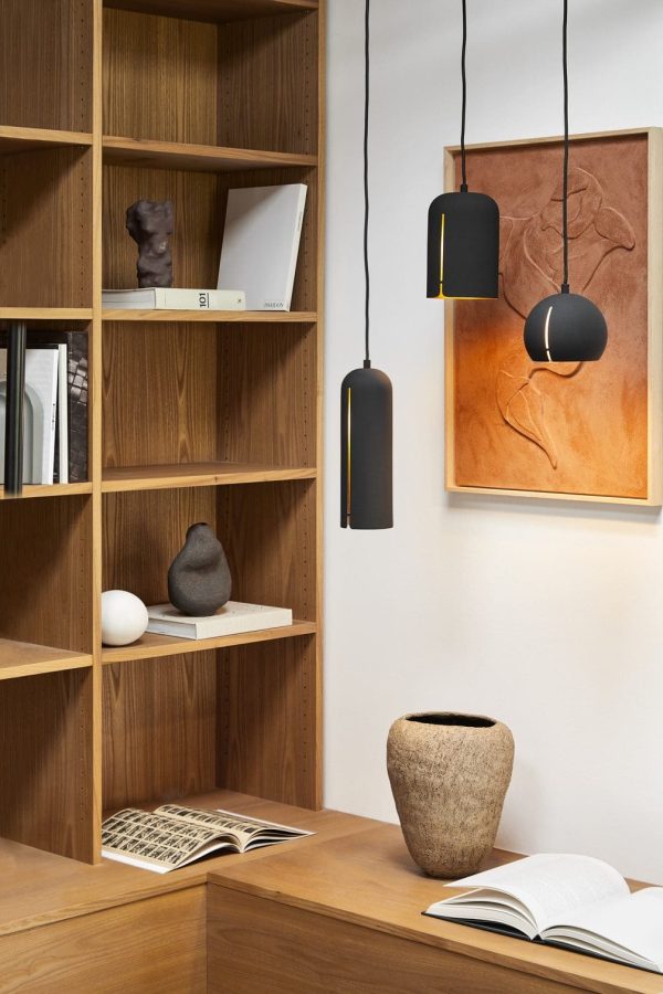 En hyggelig læsekrog med en bogreol i træ fyldt med bøger og dekorative genstande, en stor rustik vase på gulvet og tre hængende pendler. En åben bog hviler på en lille designerm