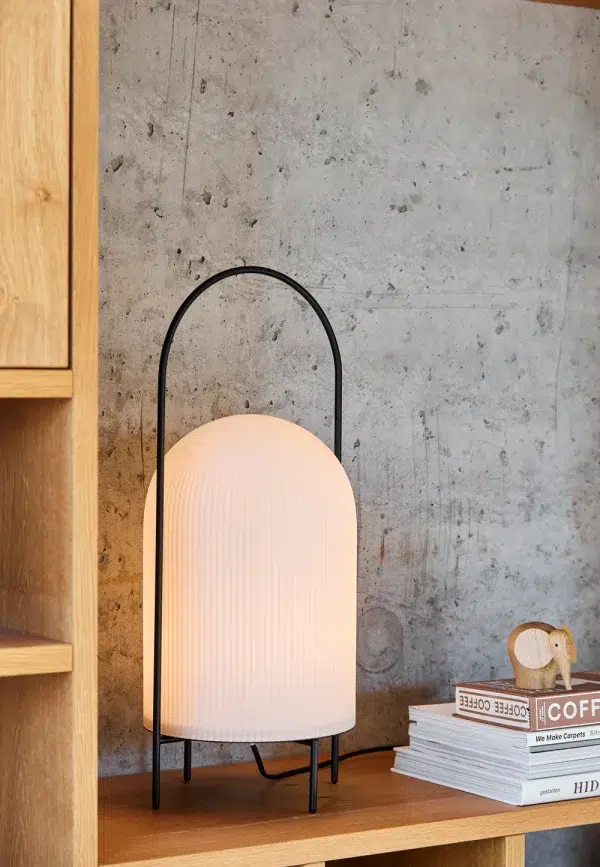 En WOUD - Ghost Bordlampe med en rillet, pastelskærm og en sort buet ramme står på en træhylde, ved siden af en stak bøger og mod en tekstureret betonvæg, der inkarnerer essensen.