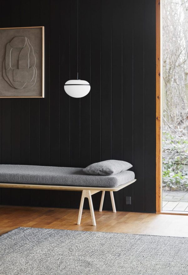 Moderne minimalistisk interiør med en smal bænk med en grå pude på en træbund mod en sort lodret panelvæg. En hvid pendel hænger over hovedet, og et abstrakt Tomorrows-design dekorerer