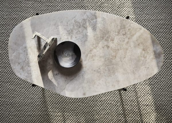 Set ovenfra og ned af et gråt marmorbord med en organisk, uregelmæssig form og et centralt hul med et slankt metalhåndtag. Denne designermøbel er placeret på en tekstureret, vævet gulvmåtte.
