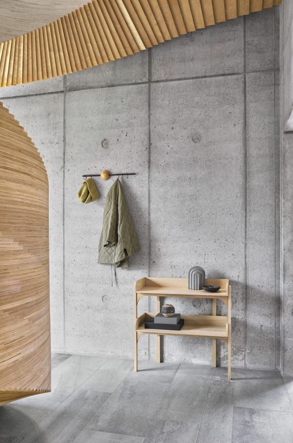 Moderne designermøbel interiørdesign med en betonvæg med træaccenter, en bænk med sko og bøger under, og en frakke og hat, der hænger ovenover på minimalistiske kroge.