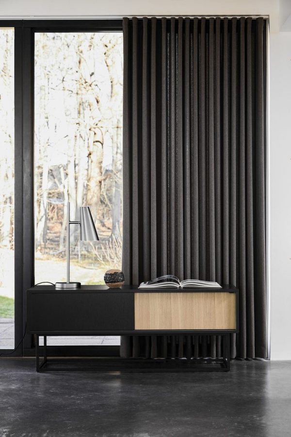 En moderne læsekrog med et stort vindue med sorte gardiner i fuld længde, et sort sidebord med bøger og en lampe og en designertræbænk i et værelse med et poleret betongulv.