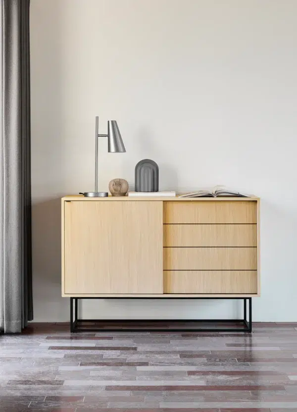 Et minimalistisk designermøbelskab med to skuffer og en lampe på toppen, placeret mod en almindelig væg med gråt gulv. En bog, små skulpturer og en billedramme pryder også skabet