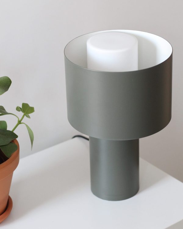 En moderne grå cylindrisk lampe med en semi-transparent skærm, placeret på et hvidt bord ved siden af en terracotta krukke med en lille grøn plante fra Tomorrowsdesign.