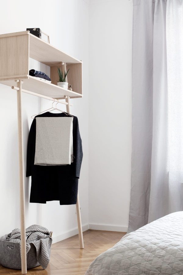 Et minimalistisk soveværelseshjørne med en WOUD - Töjbox Garderobe med en sort og grå sweater, en hylde over med sko og bøger og et gråt sengetæppe. Naturligt lys filtrerer gennem et hvidt gardin.