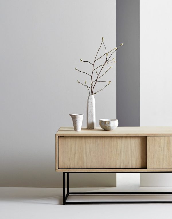 Et minimalistisk interiørdesign med en lys træskænk med metalramme, dekoreret med en høj grå vase med sarte grene, flankeret af to mindre metalliske skåle. En neutral-farvet væg fungerer som