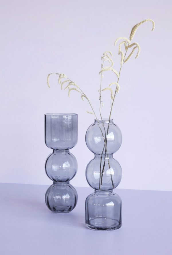 En moderne HÜBSCH - Circle Vase (sæt af 2) med tre stablede kugleformede sektioner af gennemsigtigt glas, indeholdende tørrede prydgræsser med krøllede spidser, mod en lys lilla baggrund.