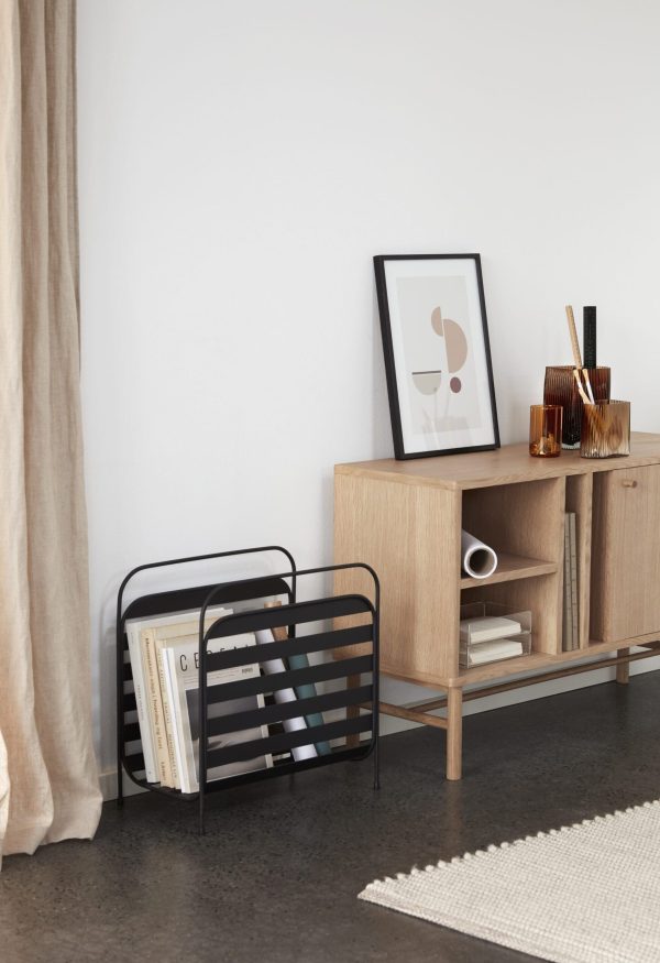 Et moderne stuehjørne med en opbevaringskonsol i træ, et indrammet abstrakt kunstværk af Tomorrowsdesign, en magasinstativ med bøger, dekorative genstande og et beige gardin. Gulvet er gråt,