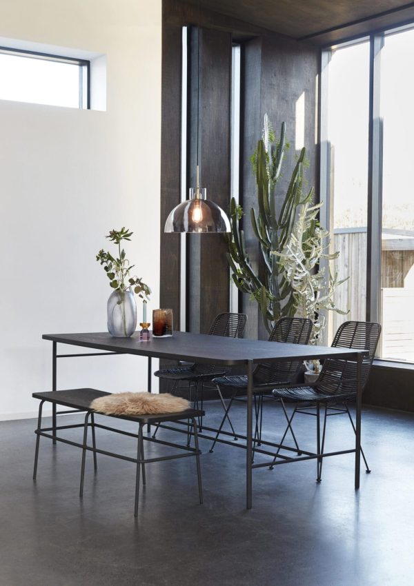 En moderne spiseplads med et minimalistisk sort bord og stole, en stor kaktus i hjørnet og en metallisk pendel hængende over hovedet. Rummet har slanke betongulve, store vinduer og er