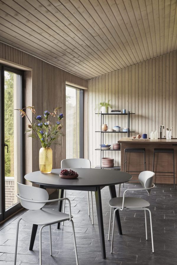 Moderne køkkeninteriør med træspaltevæg og -loft, med et cirkulært gråt bord med hvide stole, store vinduer og en hylde med keramik. Dette rum følger Tomorrowsdesigns etos