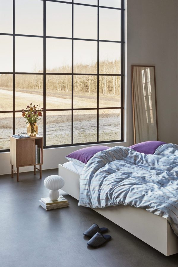 Et moderne soveværelse med HÜBSCH - Solace Sengetøj med en lav seng med blåt og hvidt sengetøj, et sidebord i træ med vase med blomster, store gulv-til-loft vinduer med udsigt over et goldt landskab og minimal indretning.