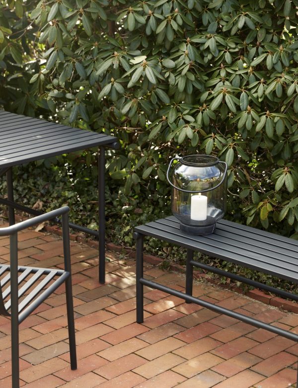 Et hyggeligt udendørs opholdsområde med to mørke metalbænke på en muret terrasse, omgivet af frodige grønne buske. En glaslanterne med hvidt lys er placeret på en af designermøbelbænkene.