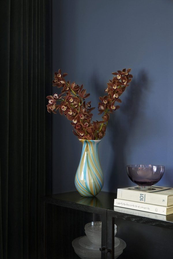 En vase med hvirvlende blå og hvide mønstre holder røde orkideer på et mørkt, elegant interiørbord mod en marineblå væg, ledsaget af en stak bøger og pyntegenstande.