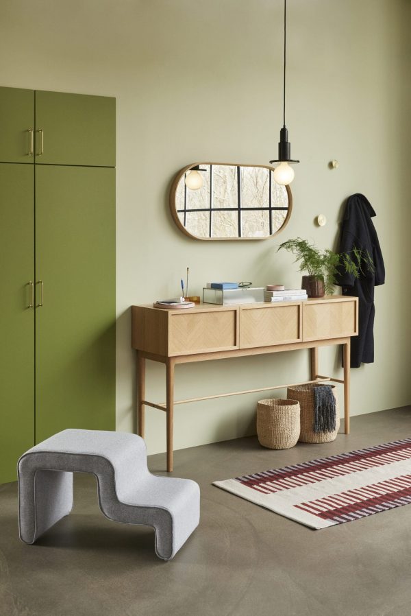 En moderne entré med et konsolbord i træ med små skuffer, et unikt spejl over det og en grå skulpturel bænk. Et grønt skab og hængende tøj fremhæver rummet, oplyst af et ophæng