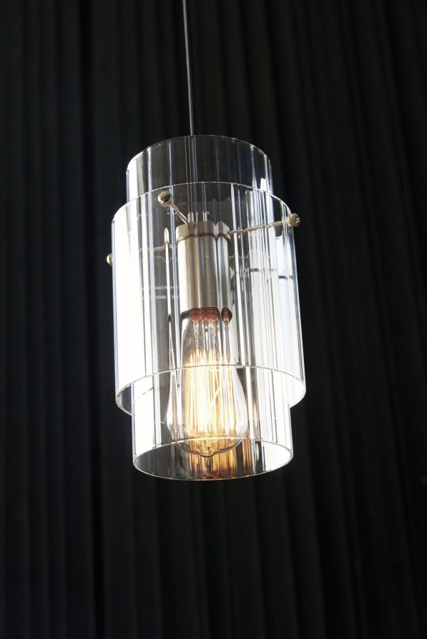 En moderne pendel, et Tomorrows-designstykke, med en klar cylindrisk glasskærm og en synlig glødetrådspære, der hænger på en mørk rillet baggrund.