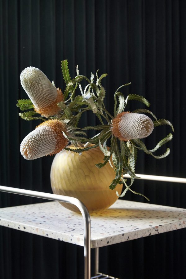 En vase med eksotiske teksturerede blomster, inklusive aflange hvide blomster, sidder på en designer-spættet bordplade mod en mørk bølget baggrund, oplyst af naturligt lys.