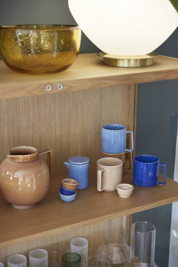En moderne, stilfuld hylde med et udvalg af keramik, inklusive blå og terracotta krus, en stor terracotta kande og forskellige glas. En stor, glødende rund lampe sidder