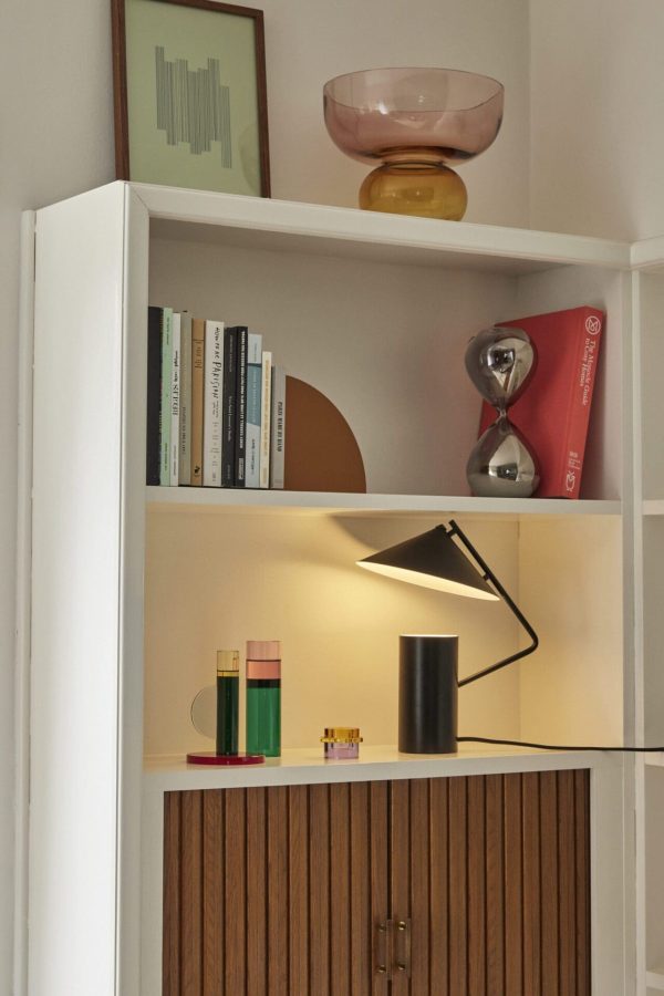 En moderne hvid designermøbel med forskellige pynteting, herunder bøger, en lampe og små farverige glasgenstande. Den øverste hylde rummer en lyserød skål og en metallisk skulptur.