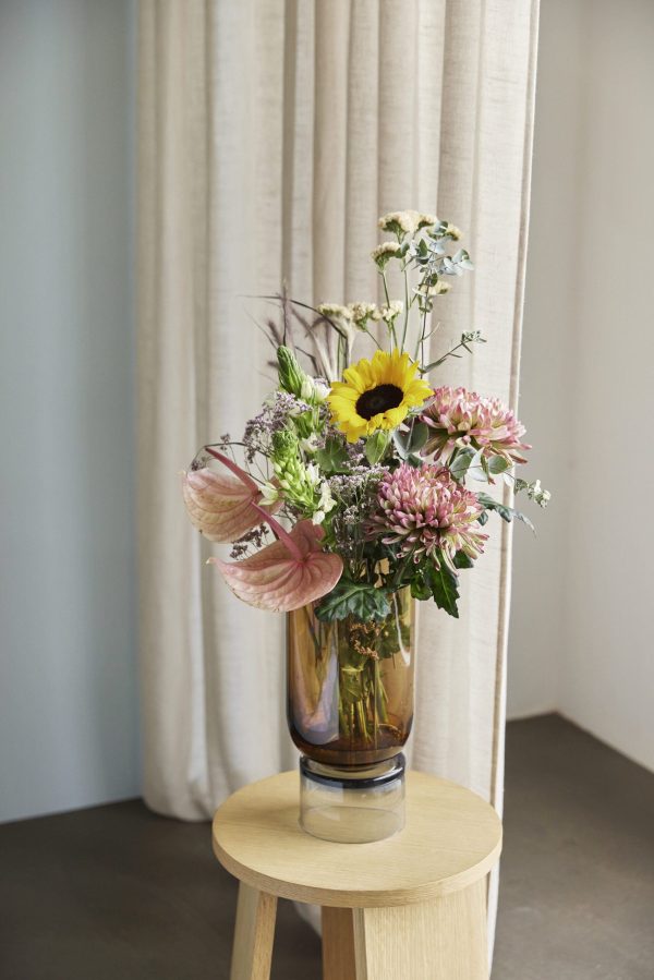 En HÜBSCH - Stand Vase Stor fyldt med en farverig buket blomster, inklusive en solsikke og lyserøde blomster, placeret på en trætaburet mod en baggrund af neutrale gardiner.