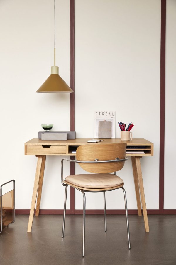 En moderne hjemmekontoropstilling med et træskrivebord med rum, parret med en HÜBSCH - Arch Spisebordsstol Hynde Natural stol. en messing pendel hænger over hovedet, og minimalistisk indretning, inklusive et magasin og brevpapirholder, pryder skrivebordet.