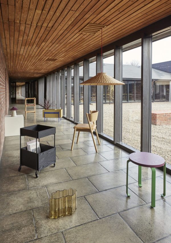 Interiør af en moderne gang med store glasvinduer, træloft og stengulv. Har minimalistiske møbler, herunder et lille bord, stole og flytbare hylder, med unikke belysningsarmaturer.