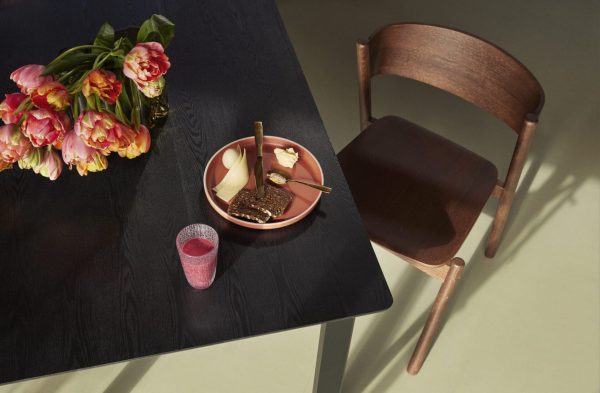En moderne spisestue med et elegant sort bord med en tulipanbuket, en skål med pærer og ost på en tallerken, ved siden af en stilfuld træmøbelstol og et blødt sollys.