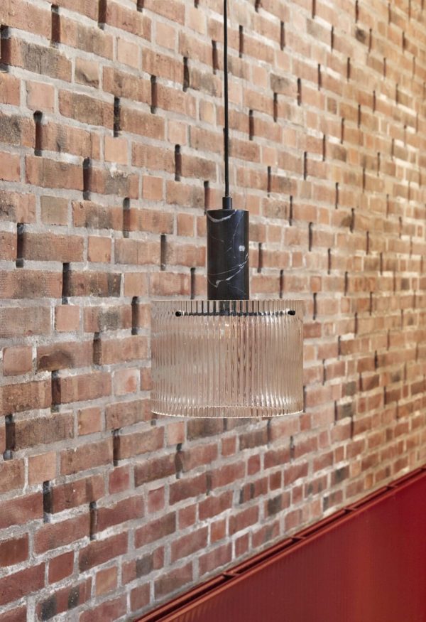 En HÜBSCH - Revolve Loftlampe Ø25 med en klar, rektangulær glasskærm hænger mod en struktureret murstensvæg, med en del af en rød overflade synlig nederst på billedet.