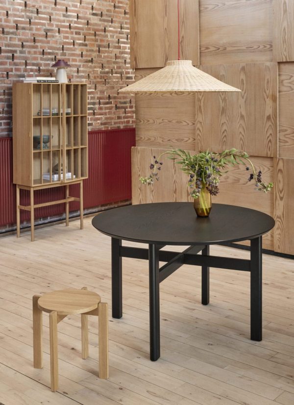Et moderne træinteriør med et rundt sort bord med en vase med blomster, en matchende skammel og et designermøbel skab med glasfront mod en baggrund af træpaneler og en rød
