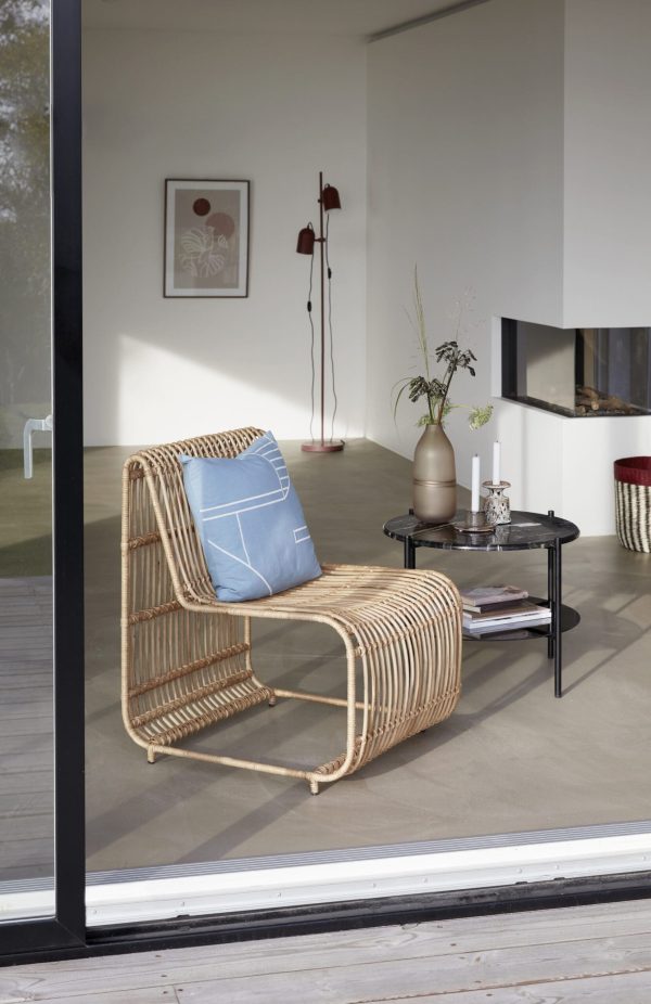 En moderne stue med en designerlænestol, blå pude, lille sort sofabord med bøger og en vase på baggrund af en stor glasdør, der fører til en terrasse.