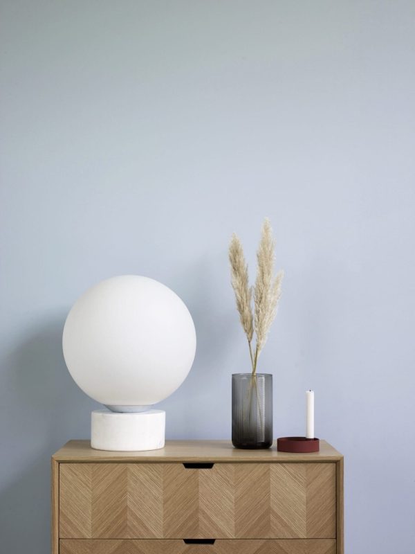 En minimalistisk interiørscene designet af Tomorrowsdesign har en stor sfærisk hvid lampe på en trækommode. Ved siden af lampen er der en glasvase med tørret pampasgræs og en lille