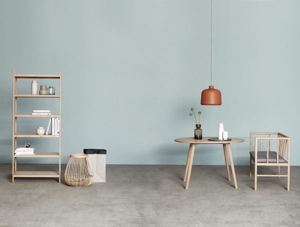 Et minimalistisk værelse med pastelblå vægge, med træmøbler: en reol, et rundt bord med stole og en HÜBSCH - Landromat Kurve pendel. Et par dekorative ting såsom bøger og vaser er pænt arrangeret.