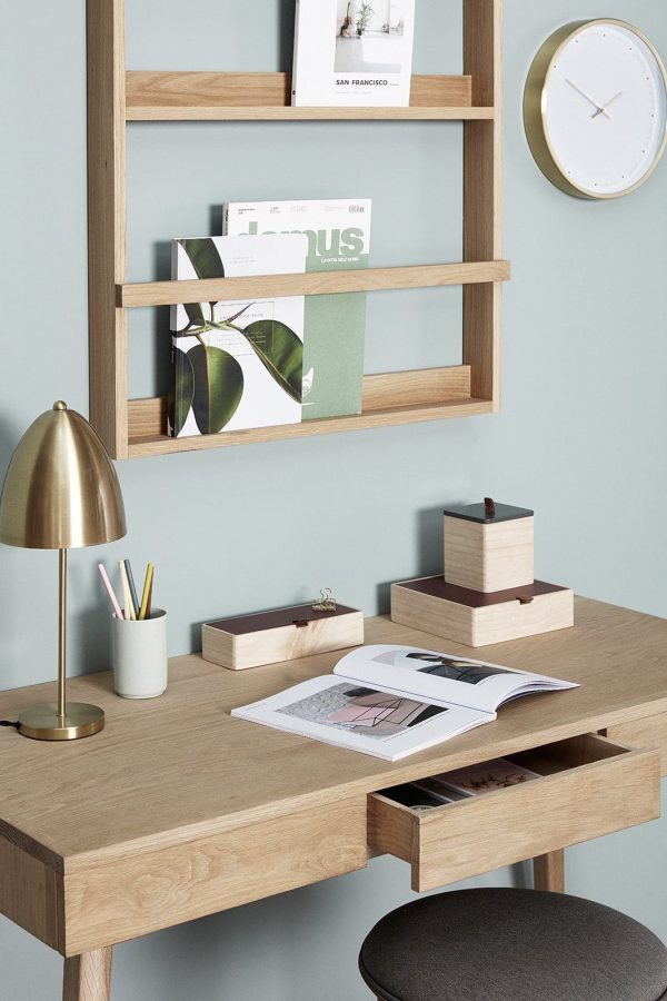 Et pænt organiseret skrivebord i træ med åbne skuffer, udstilling af bøger og et magasin. Over skrivebordet rummer en matchende træhylde bøger, et ur og en lampe. Et moderne, minimalistisk kontorsetup med