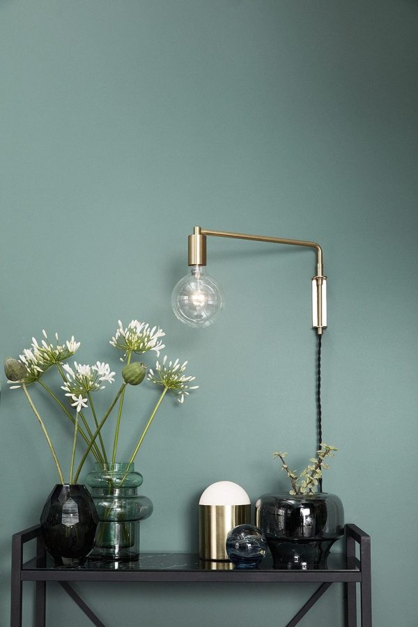 En vægmonteret messinglampe med en synlig pære strækker sig over et sort metal konsolbord mod en mørk blågrøn væg. Bordet, der viser moderne interiørdesign, rummer to vaser med hvide blomster og