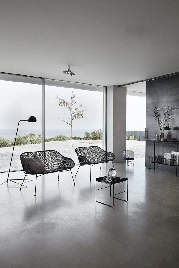 En moderne stue med store vinduer med udsigt over havet, med to sorte trådstole, et lille sort bord og minimalistisk indretning. Et ensomt træ er synligt udenfor.