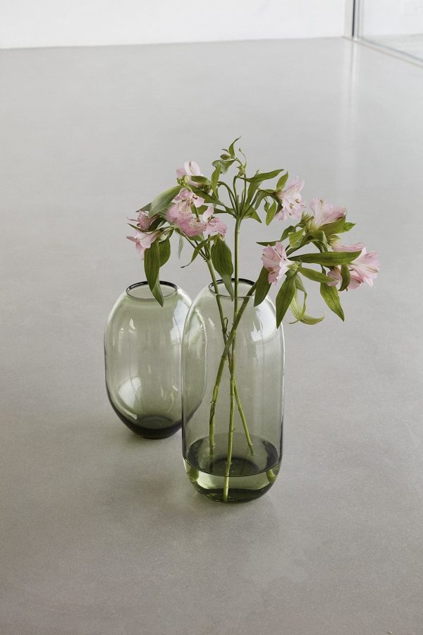 To transparente glasvaser på en grå overflade, den ene med en stilk med flere lyserøde blomster. Vaserne, som eksemplificerer enkel, men elegant indretning, har en lille mængde vand ved