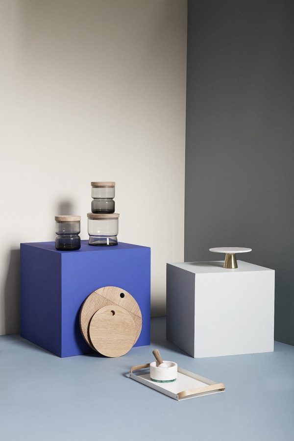 Et minimalistisk display med en blå kube med HÜBSCH - Monarch Skærebræt Rund (sæt af 2), hvide æsker og en lille metalbakke med et stearinlys. baggrunden omfatter en grå og beige væg.